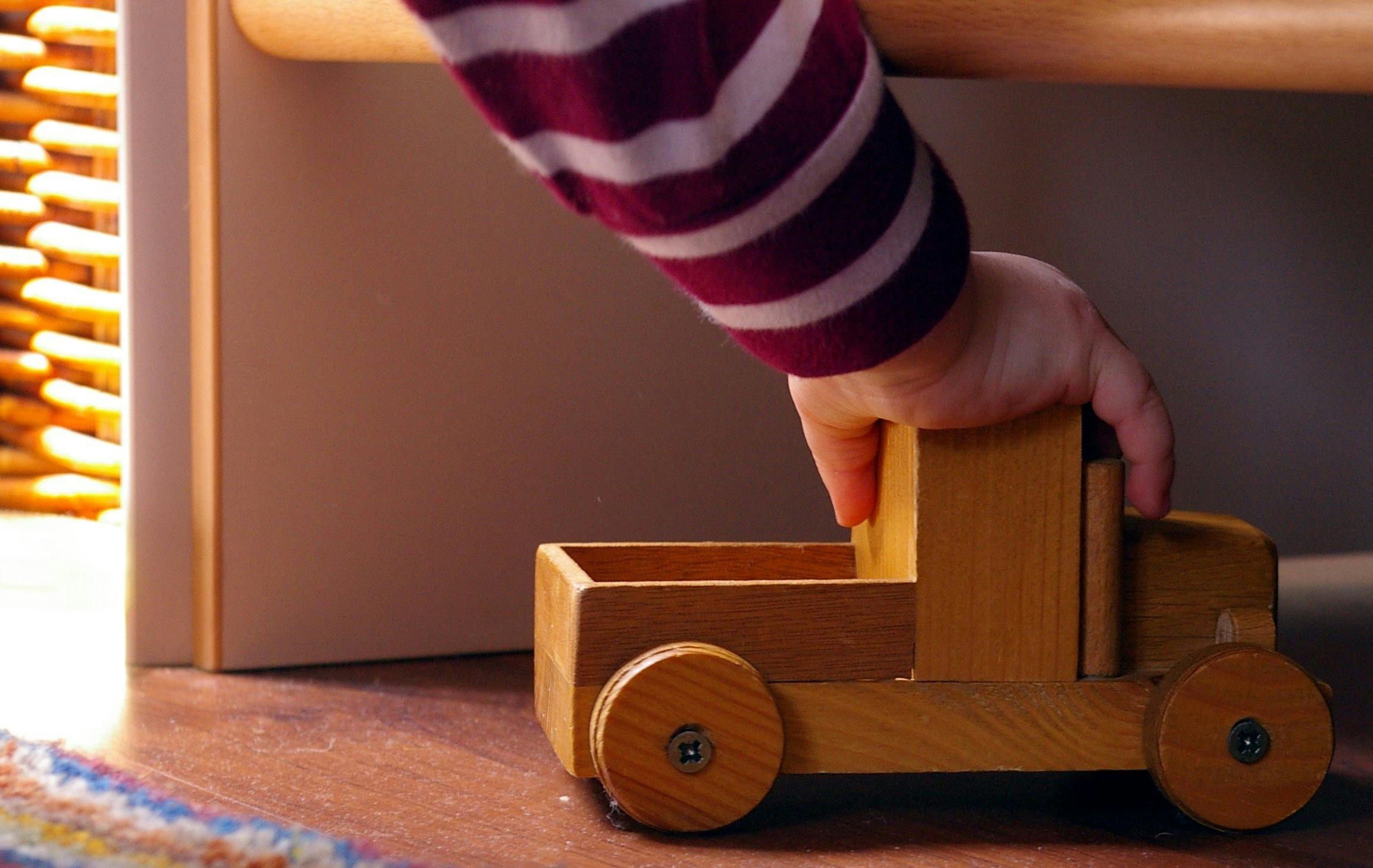 Eine Kinderhand, die ein Spielzeugauto aus Holz über den Boden fährt.